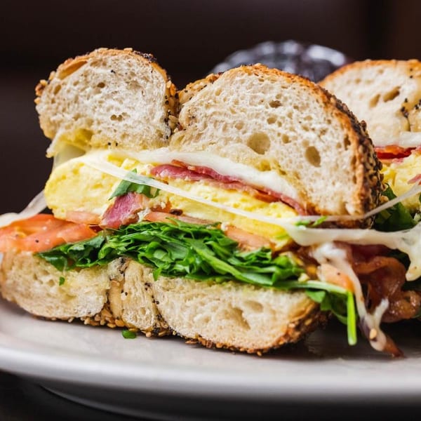 Close-up of a bagel sandwich with egg, bacon, cheese, lettuce, and tomato on a white plate.