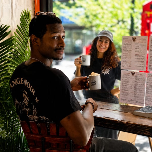Man in black Ange Noir shirt holding a mug, smiling at the camera, with a woman behind him.